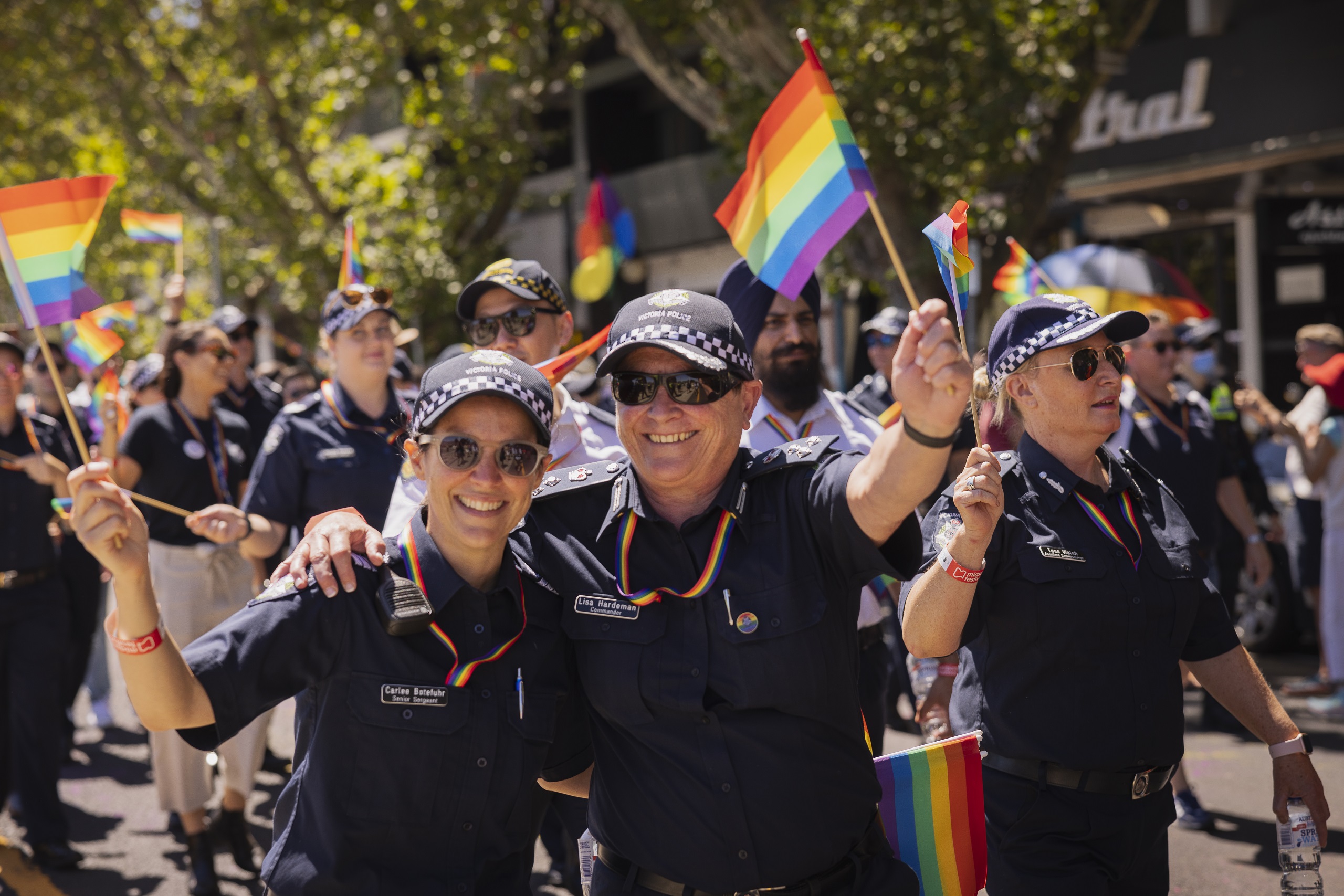Supporting family and friends Police officers walking along the road and footpath during the day in front of shops taking part in a peaceful pride march for the LGBTQIA+ community. Two police officers in uniform are smiling at the camera waving rainbow-coloured flags waving