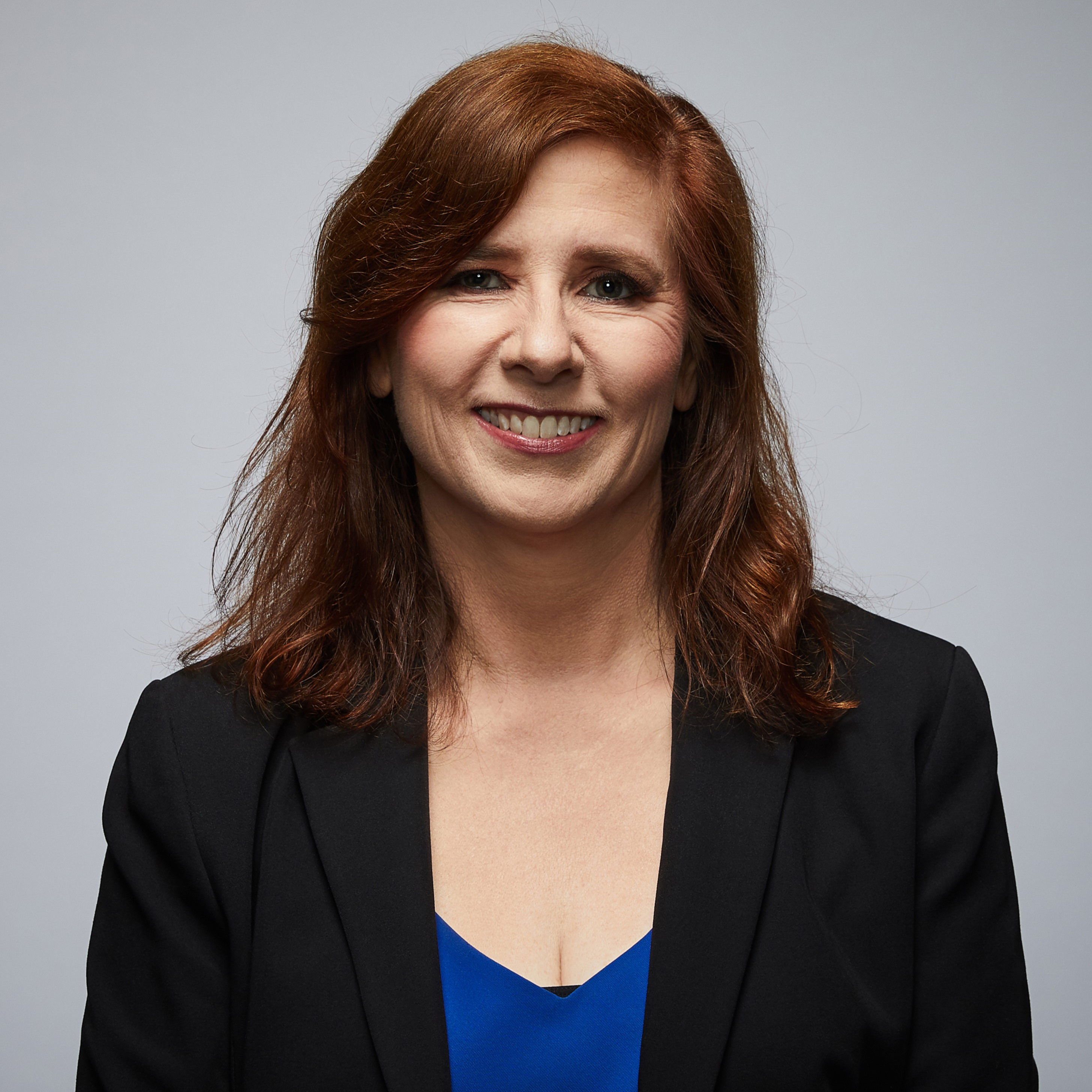 Image of police spouse looking at camera wearing a dark blue jacket or blazer. Light skin and auburn hair